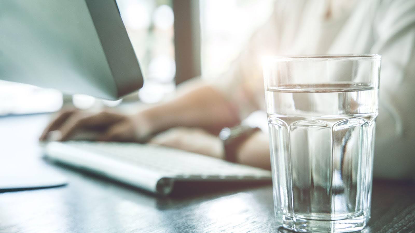 A glass of water on a desk with a person typing on a computer keyboard in the background.