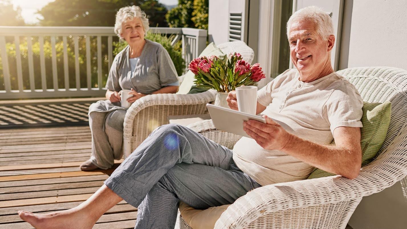 A senior couple enjoying a sunny day on their home's deck. The woman is seated in a wicker chair smiling gently while holding a book, and the man, also in a wicker chair, is using a tablet and holding a coffee cup, with a broad smile on his face.