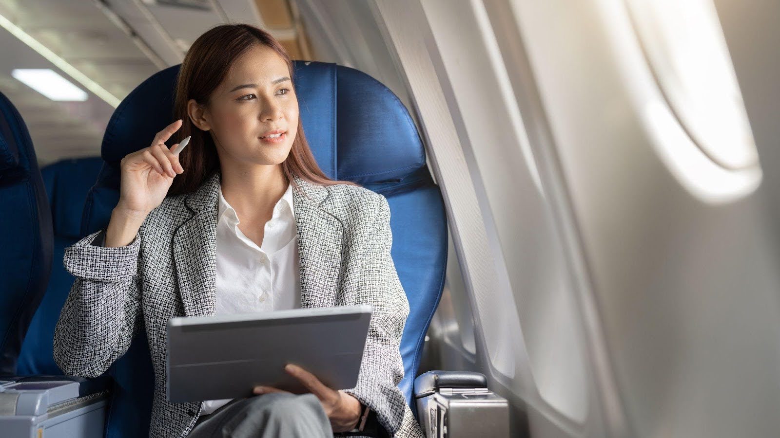 A professional woman on a long-haul flight, thoughtfully engaged with a digital tablet while looking out the aeroplane window. 