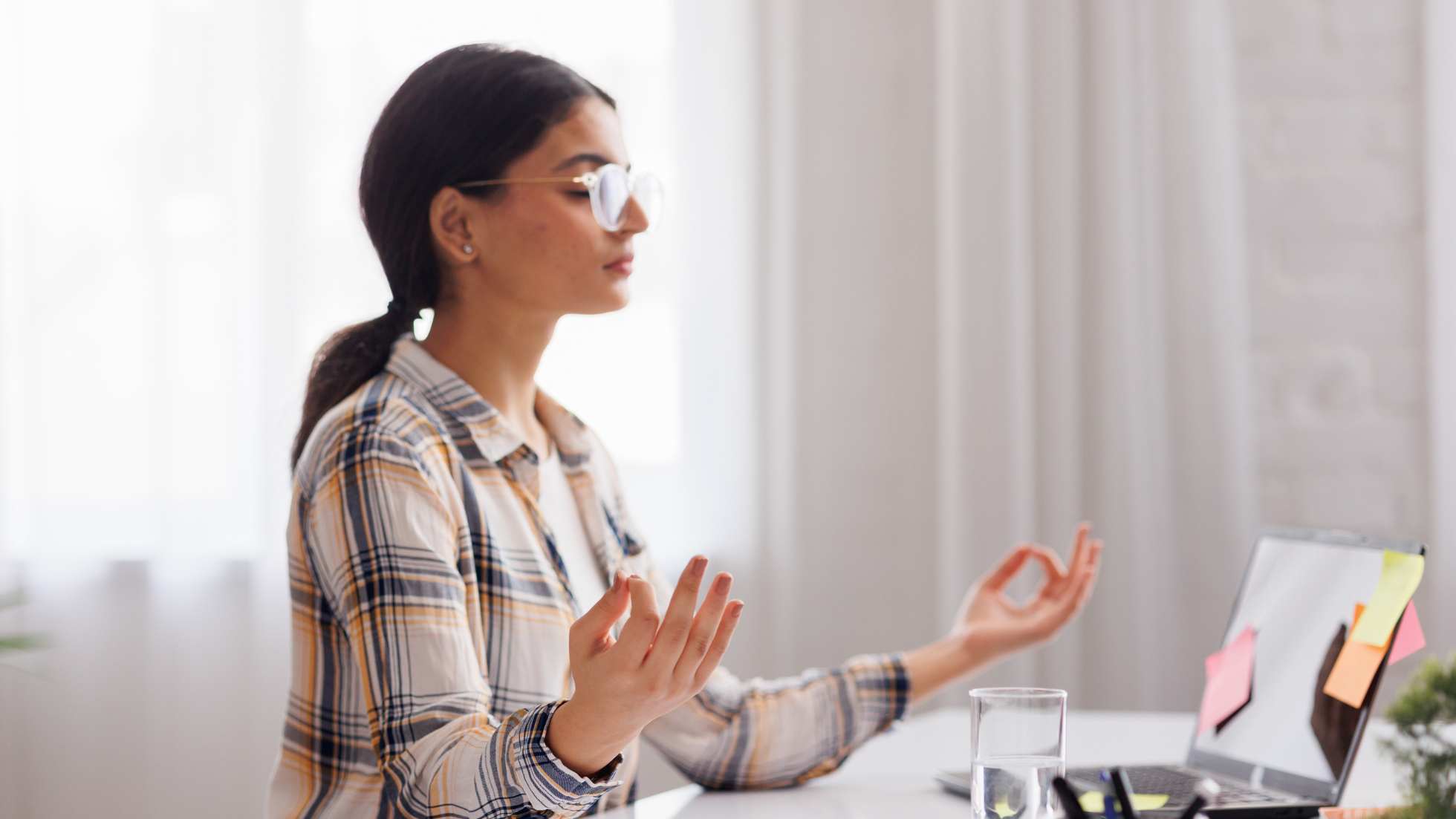 A young woman with glasses, wearing a plaid shirt, sits at her desk practicing meditation with her eyes closed and hands in a mudra position. She is in a bright, modern office setting, with a laptop adorned with sticky notes in front of her, suggesting a moment of mindfulness during a busy workday.